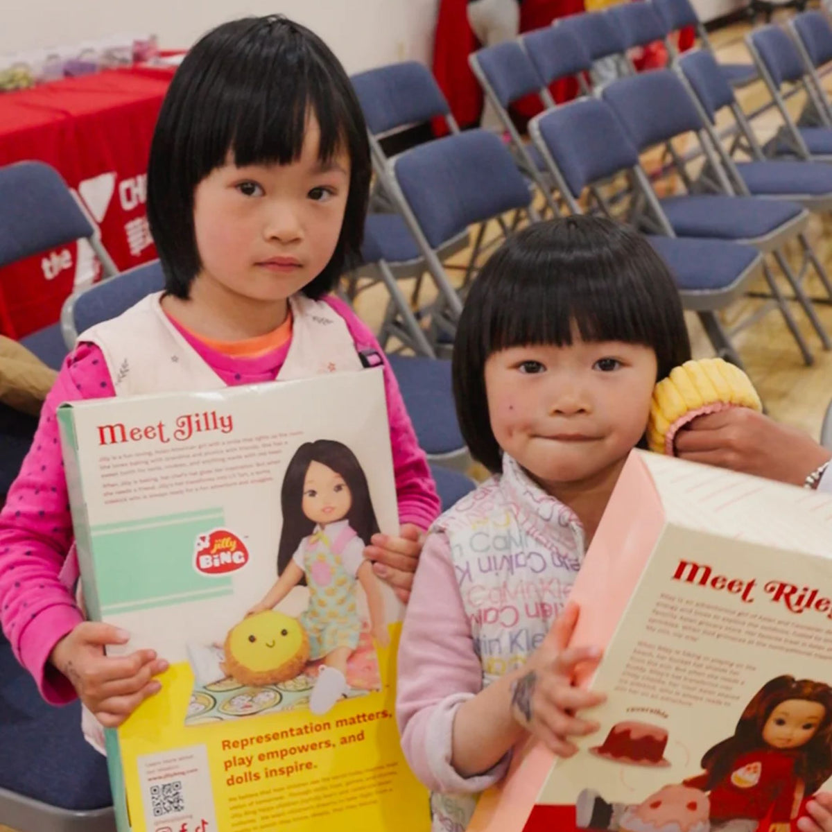 Two Asian American girls holding Jilly Bing 14-inch doll boxes featuring Jilly and Riley at a community doll donation event.