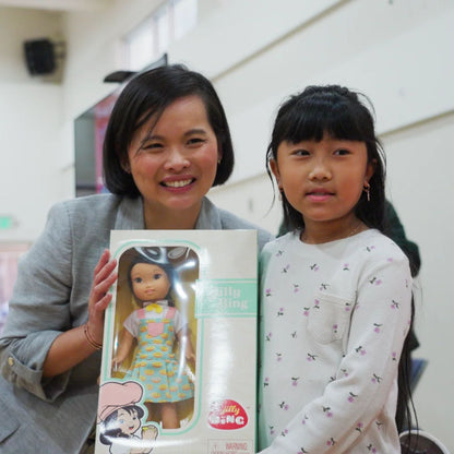 Asian American woman and young girl smiling together while holding a Jilly Bing 14-inch Asian American doll in its box at a community event.