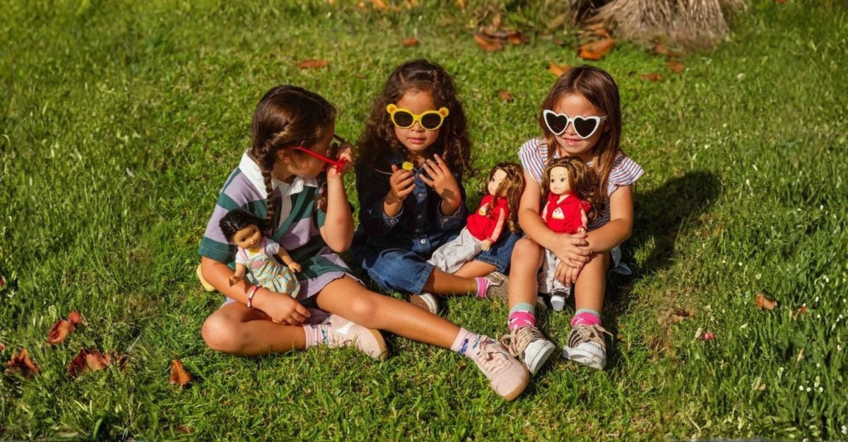 Three young girls wearing sunglasses sitting on the grass outdoors and playing with their Jilly Bing 14-inch dolls.